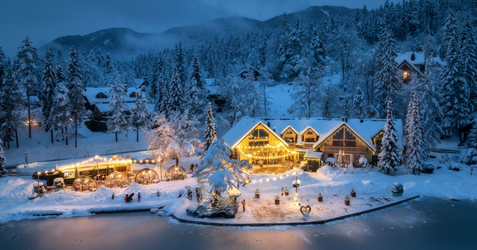 A cozy, snow-covered lakeside cabin lit up with warm lights in a winter forest setting, with surrounding pine trees and a partially frozen lake in the foreground.