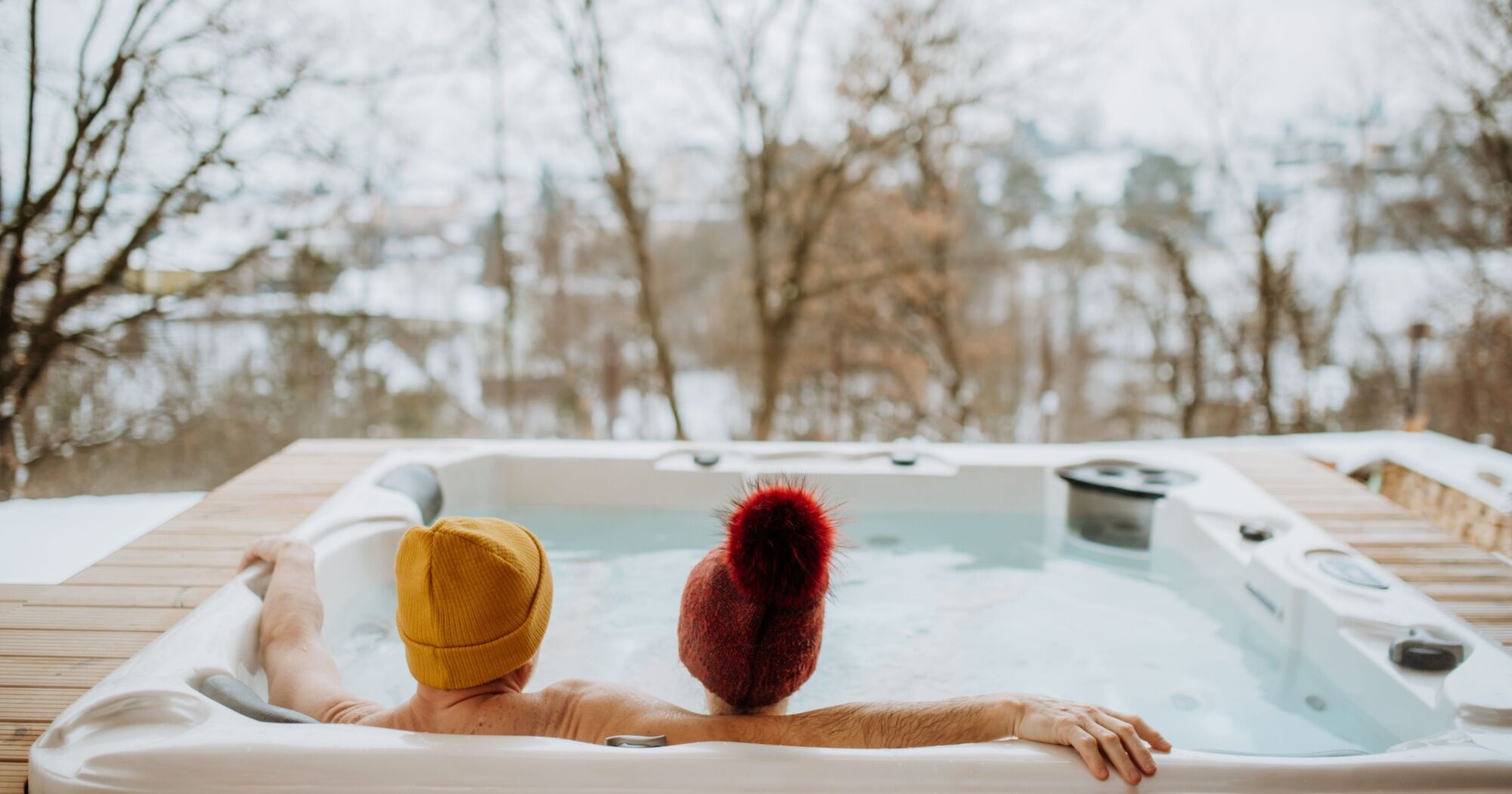 A couple relaxing in an outdoor hot tub on a snowy winter day, surrounded by a cozy wooden patio and a serene view of bare trees and a snow-covered landscape.