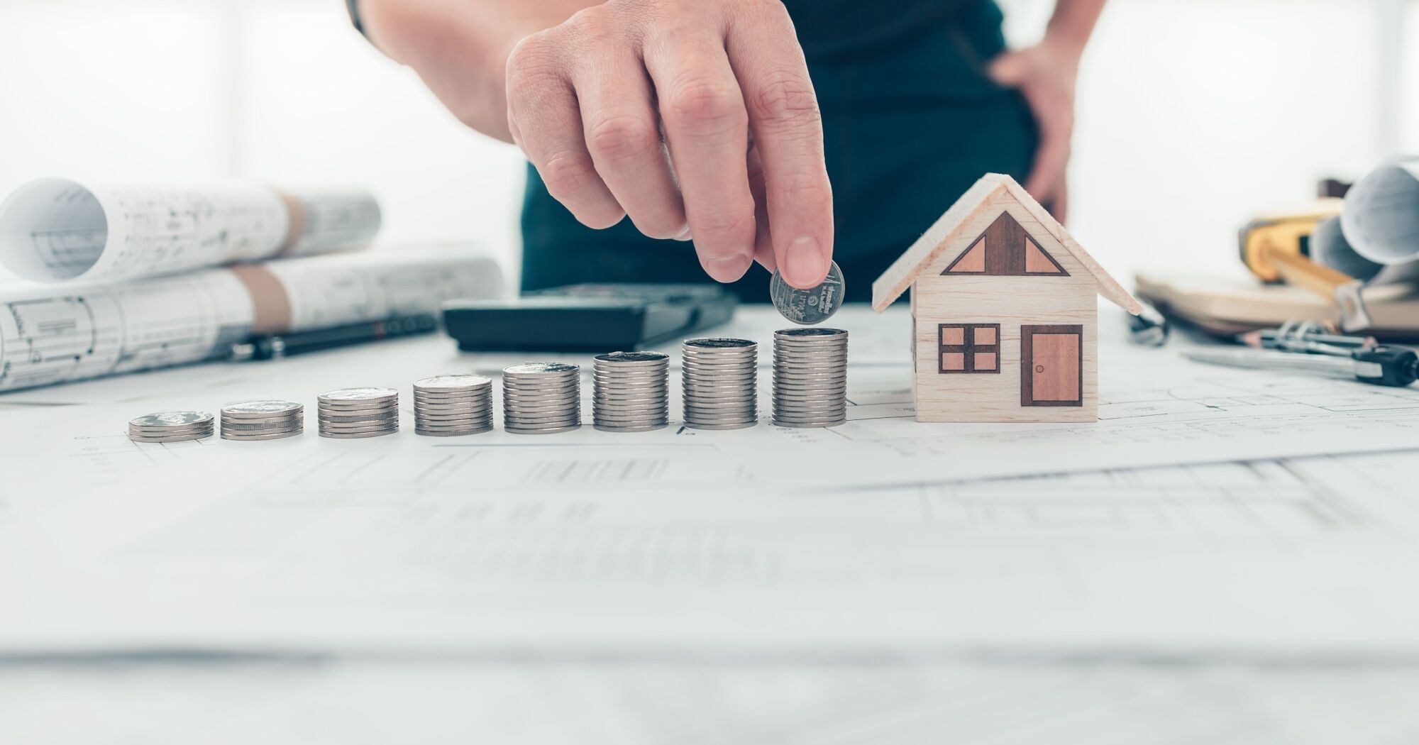 A stack of coins that resemble steps are leading up to a small wood model of a house. Underneath the house and coins are the blue prints for a home. A calculator and pen can also be seen in the background.