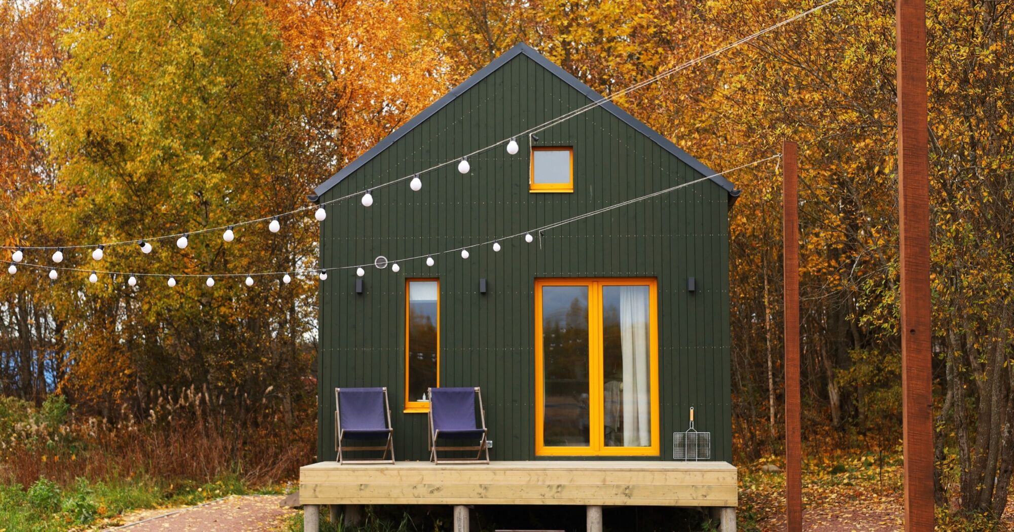 A modern dark green tiny house with bright yellow trim, string lights hanging across the front, and two lounge chairs on a small porch, surrounded by autumn trees.